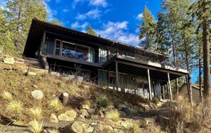 A modern two-story house with a balcony, surrounded by trees and a rocky hillside.