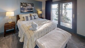 Un dormitorio con una cama grande, dos mesitas de noche con lámparas, vista a un lago a través de un ventanal, y un cuadro en la pared.