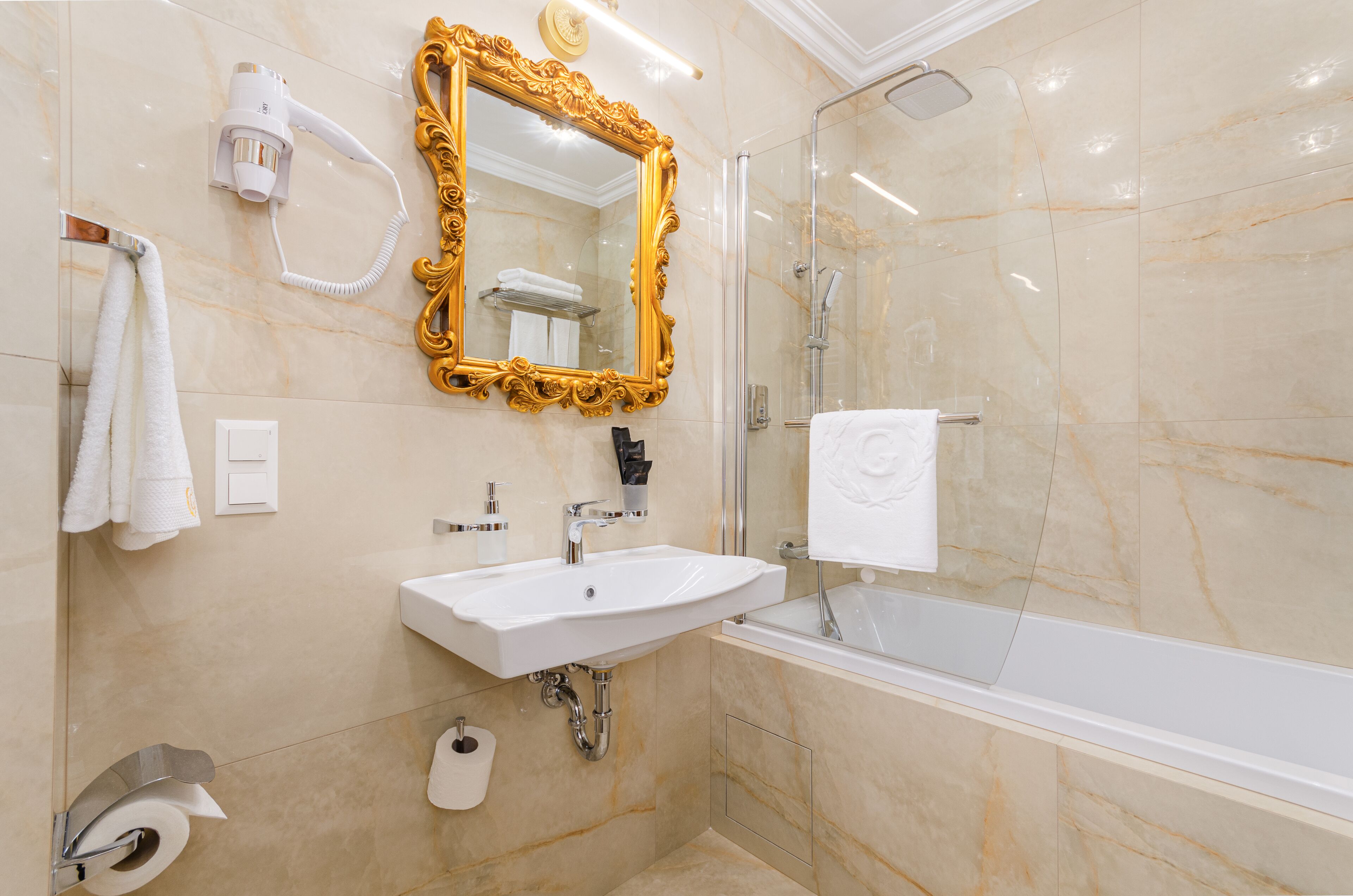 Een badkamer met een grote spiegel met gouden lijst, een witte wastafel, een douche met glazen wanden en een wit ligbad.