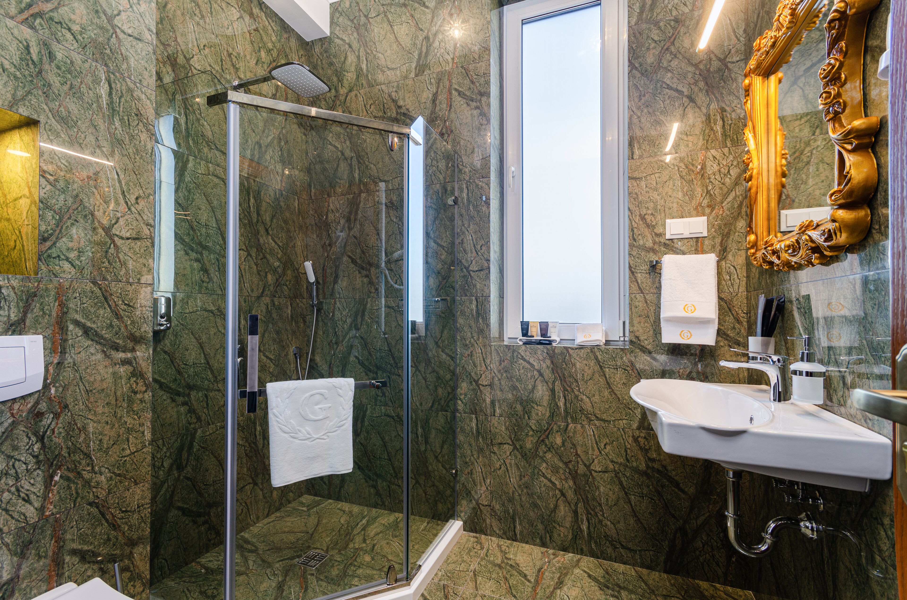 A bathroom with a glass-enclosed shower, a marble wall, a sink, and a large ornate mirror.