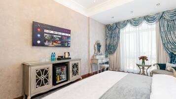 A hotel room with a large flat-screen TV displaying streaming services, a bed with a grey duvet, a vanity with a mirror, and a window with blue curtains.