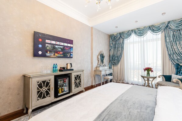 A hotel room with a large flat-screen TV displaying streaming services, a bed with a grey duvet, a vanity with a mirror, and a window with blue curtains.