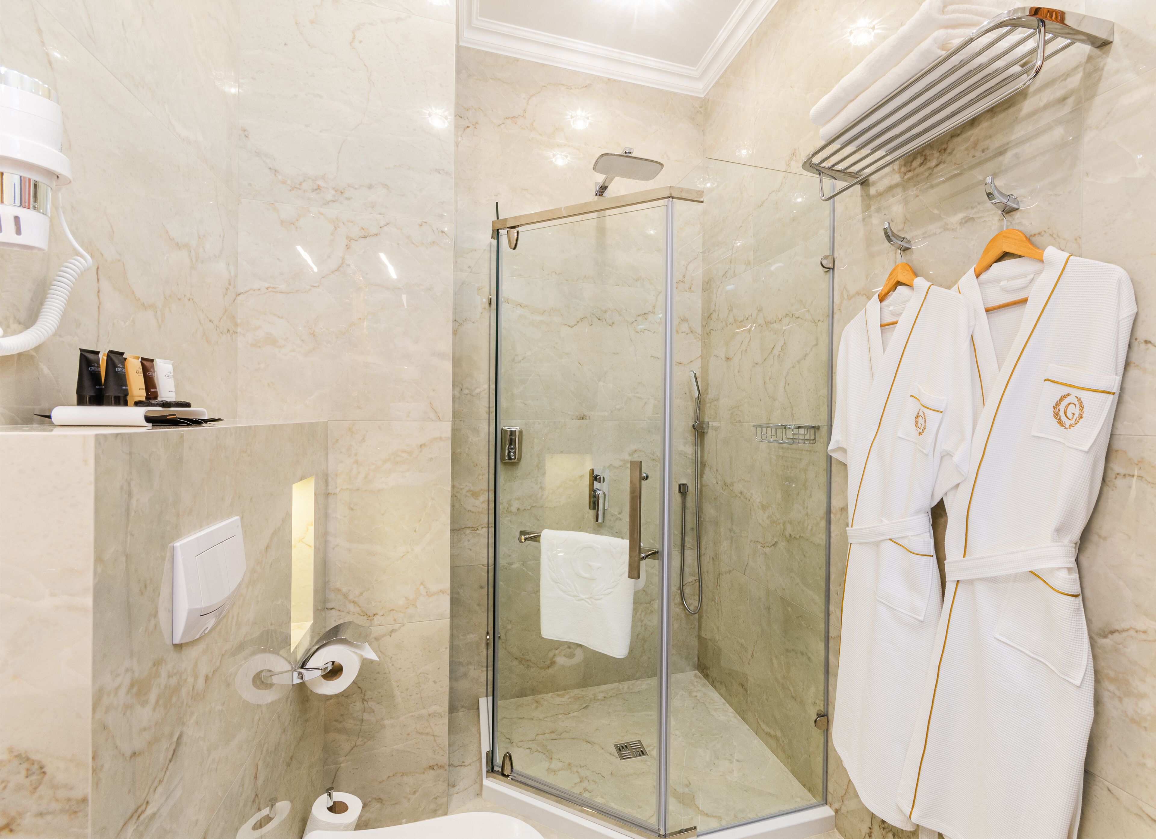 Een moderne badkamer met een douchecabine van glas, marmeren wanden, een handdoekrek met badjassen en toiletartikelen op een plank.