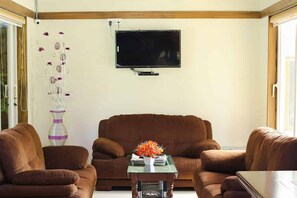 A living room with a brown sofa, a glass coffee table, and a flat-screen TV mounted on the wall. -  Leisure Inn Resort by NHospitality (Cheyyur)
