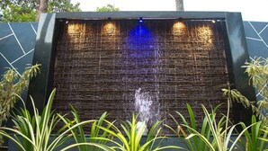 A water feature with a textured wall and a central water jet, surrounded by green plants. -  Leisure Inn Resort by NHospitality (Cheyyur)