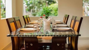 A dining table with chairs, a vase with flowers, and a window with a view of greenery. -  Leisure Inn Resort by NHospitality (Cheyyur)