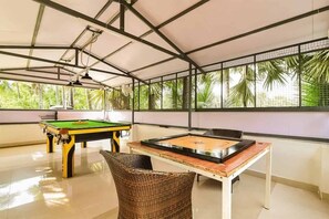 A pool table with a green felt surface, a billiards cue, and a wicker chair in a room with a high ceiling and large windows.