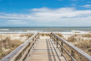 Een houten looppad naar een strand, met zand en gras aan beide kanten.