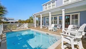 Uma área de piscina com deck de madeira, espreguiçadeiras brancas e um espaço de estar coberto com janelas.