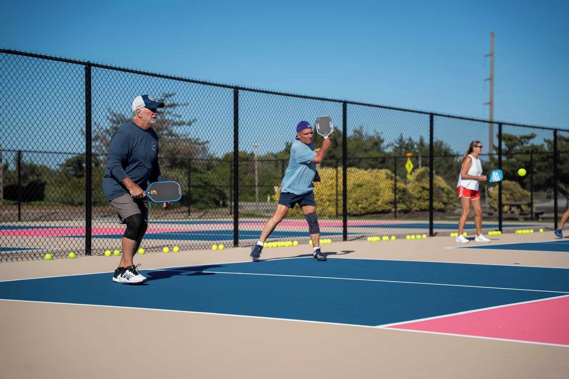 En tennisbana med tre spelare, ett stängsel av sammanlänkade metallkedjor och en klar himmel.