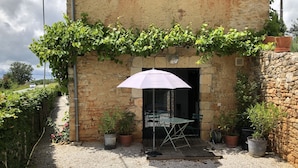 Un bâtiment rustique en pierre, doté d’une terrasse en gravier, d’un parasol blanc et de plantes en pot.