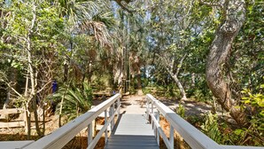 Un puente de madera que conduce a un área boscosa, con una mesa de picnic y un contenedor azul a la izquierda.
