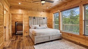 Une chambre dans une cabane en bois, avec un lit, un ventilateur de plafond et de grandes fenêtres.