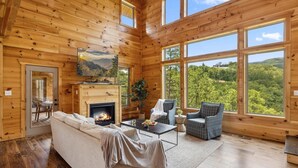 L'intérieur d'une cabane en rondins, avec une cheminée, un canapé, deux fauteuils et de grandes fenêtres donnant sur la forêt.