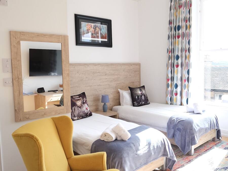 A hotel room with two beds, a large mirror, a television, a yellow armchair, and a colorful curtain.