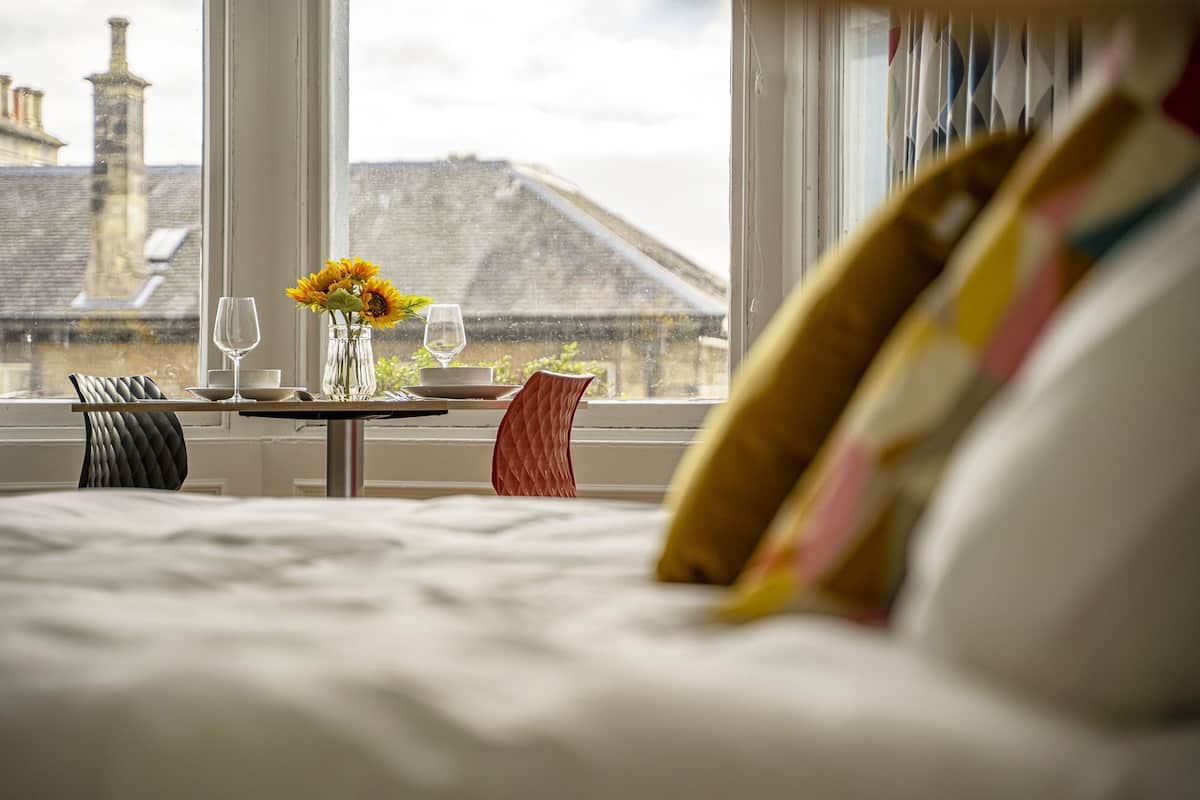 A hotel room with a bed, a dining table with a vase of flowers, and a view of buildings outside.