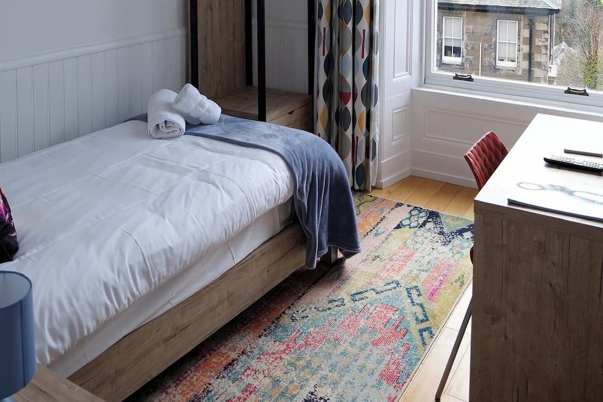 A bedroom with a bed, a desk, a window with curtains, a framed picture, and a wardrobe.