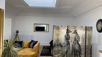 Een kamer met een gele bank, een groot schilderij, een decoratief roomdivider en een klok aan de muur.