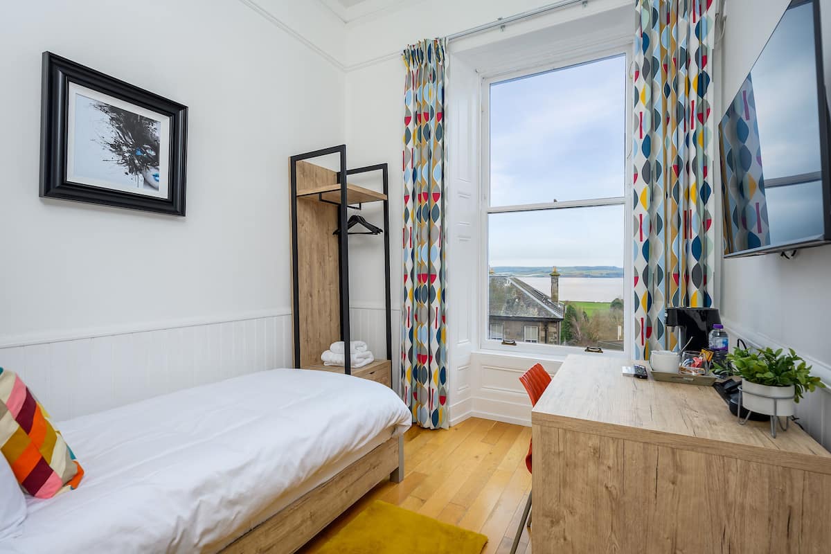 A bedroom with a bed, a wooden desk, a hanging chair, a window with curtains, and a framed picture on the wall.