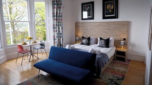 A bedroom with a bed, a blue sofa, a desk with a chair, and two bedside tables with lamps. - The Clarendon (Dundee)
