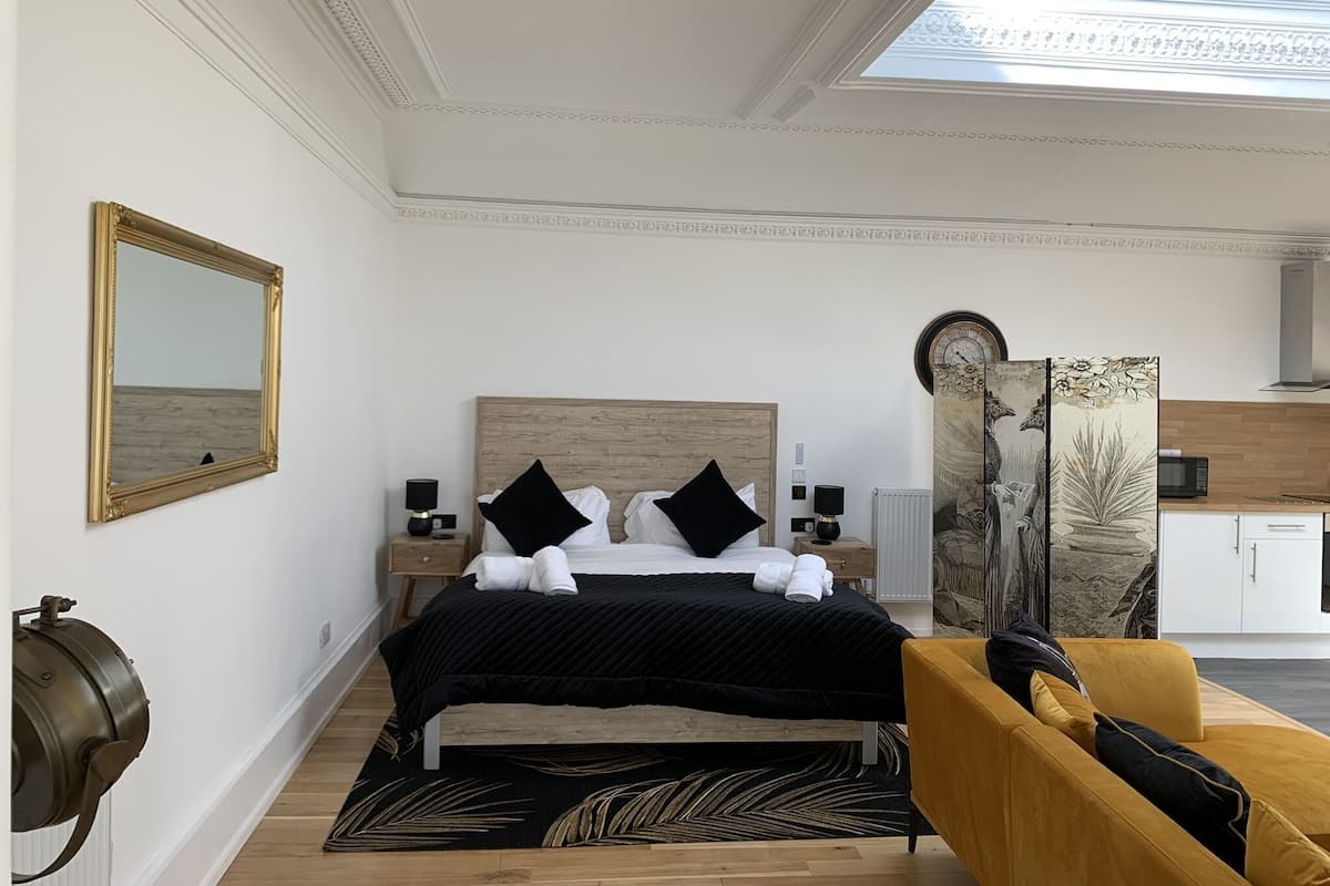 A modern hotel room with a bed, a sofa, a large mirror, a clock, and a view of the sky.