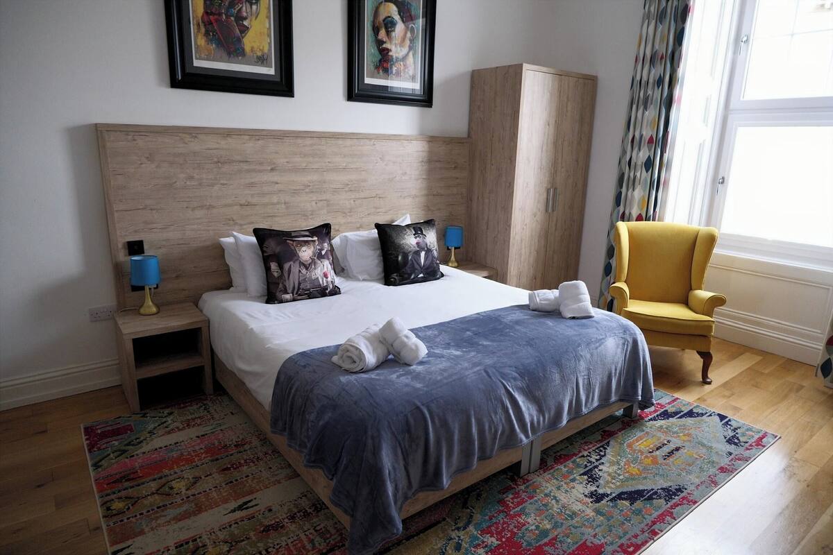 A neatly made bed with pillows, a nightstand with a lamp, a chair, a wardrobe, and a window with curtains.