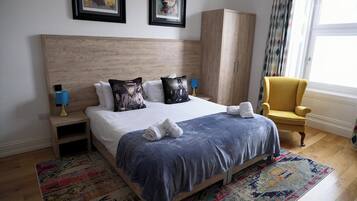 A neatly made bed with pillows, a nightstand with a lamp, a chair, a wardrobe, and a window with curtains.