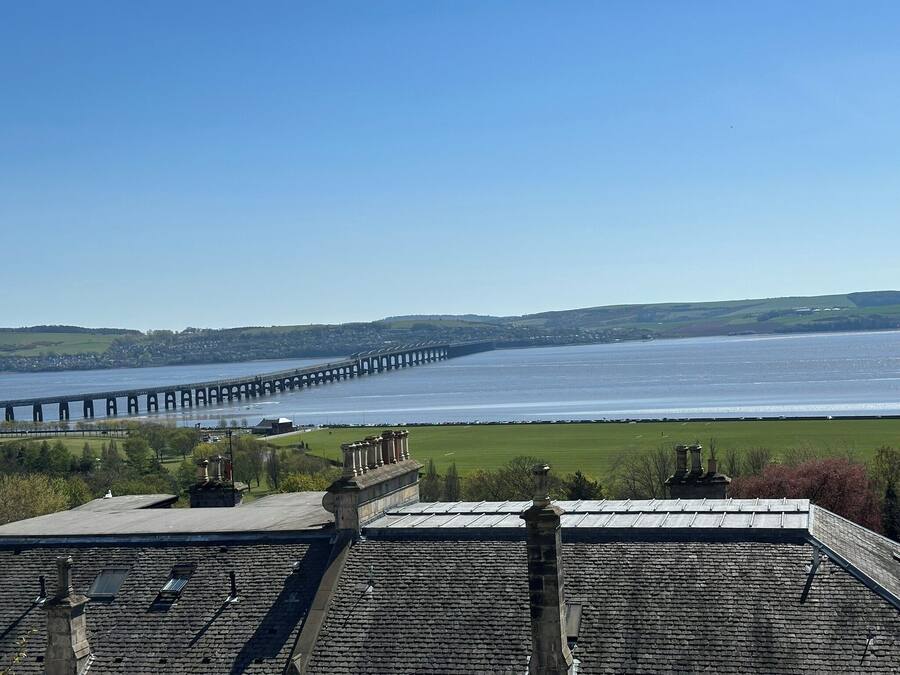 A view of a river with a long bridge, green fields, and a building with a stone roof.