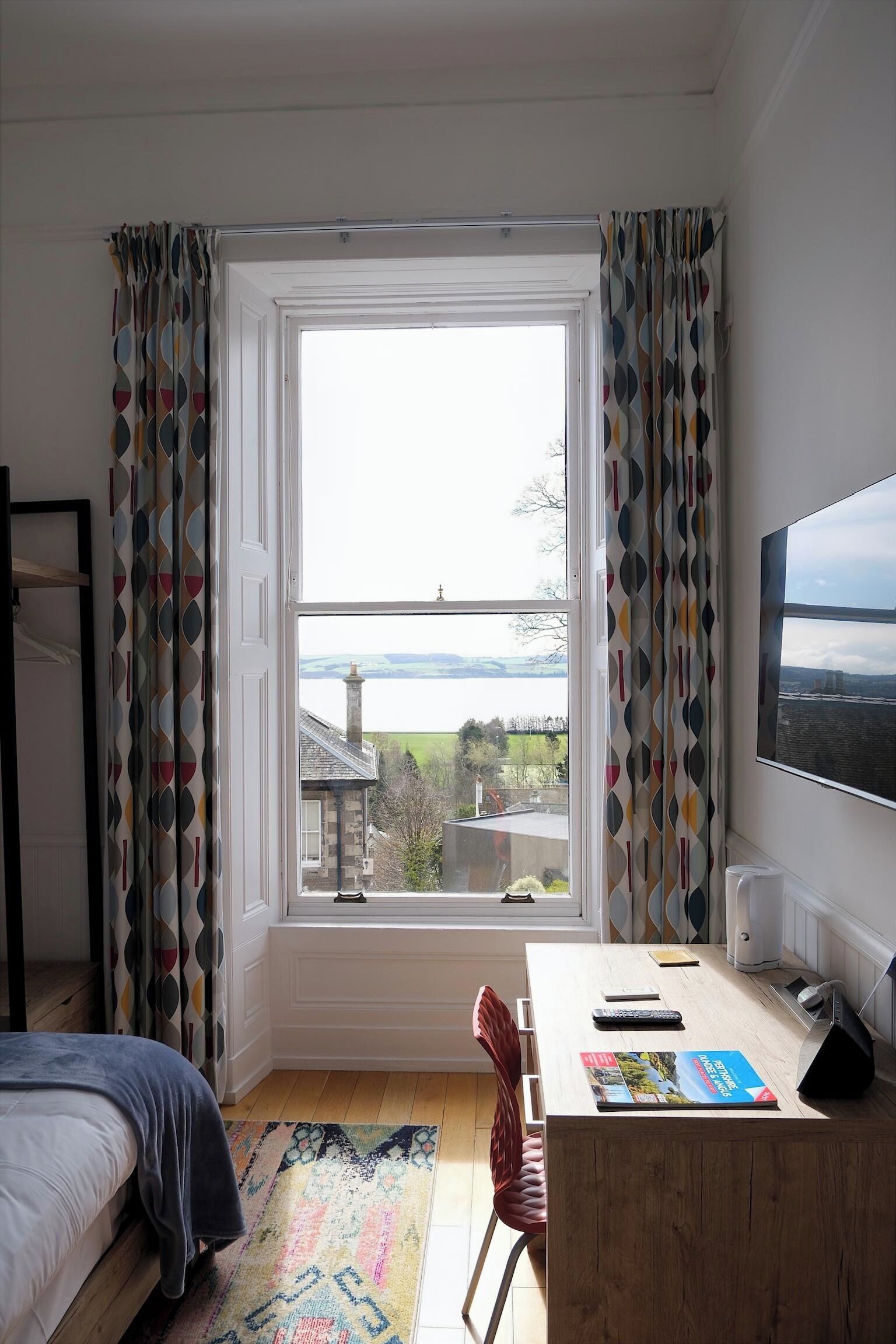 A bedroom with a large window, a bed, a desk, and a chair.