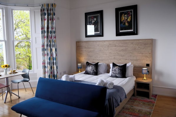 A hotel room with a bed, two framed artworks, a desk with a chair, and a sofa. - The Clarendon (Dundee)
