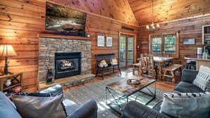 A cozy cabin interior with a stone fireplace, wooden walls, a large TV, and a dining area. - Book for fall now! Enjoy our hot tub and deck! (Gatlinburg)