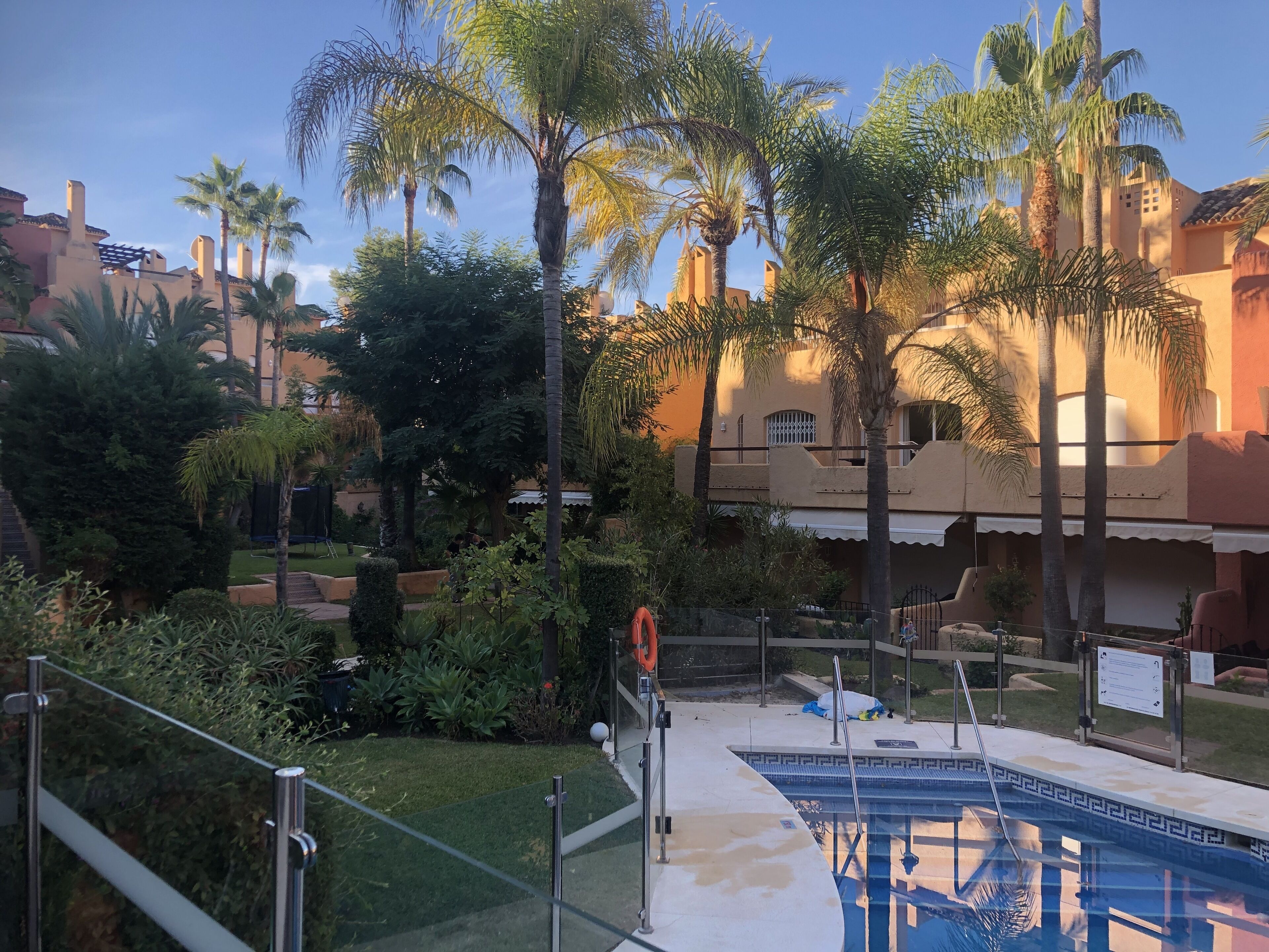 A pool area with a glass railing, palm trees, and a building with a tiled roof.