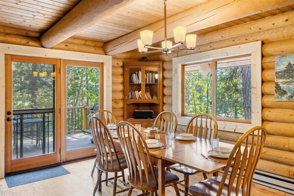 Interior de una cabaña de madera con comedor, estanterías y ventanas amplias.