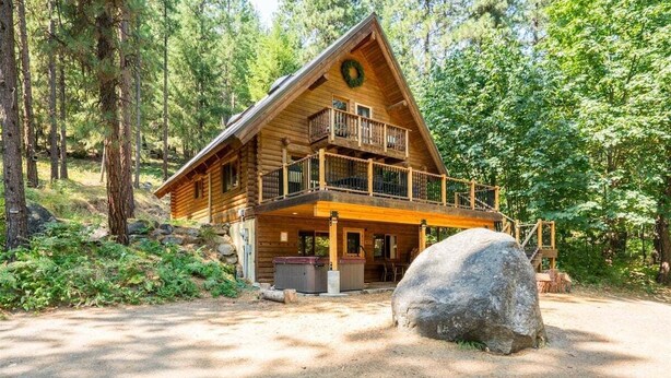 A log cabin with a balcony, a large rock in front, and a forested backdrop. - Custom Log Cabin with Great Amenities! (Leavenworth)