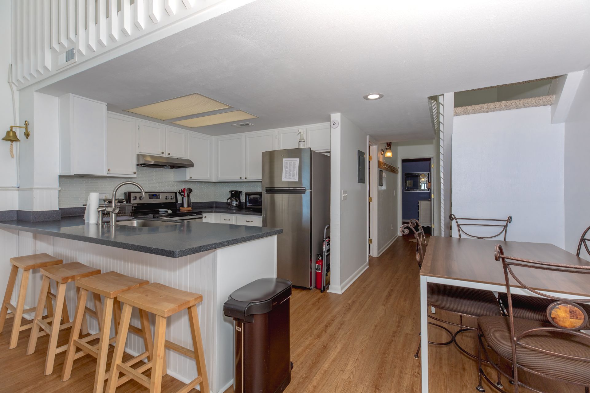 A modern kitchen with white cabinetry, a stainless steel refrigerator, a built-in oven, and a granite countertop. There are wooden stools and a trash bin.