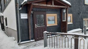 Une entrée en bois surmontée d’un toit enneigé, une enseigne indiquant qu’il s’agit d’un « Fernwohnung » (logement de vacances), et une rambarde en métal à l’avant.
