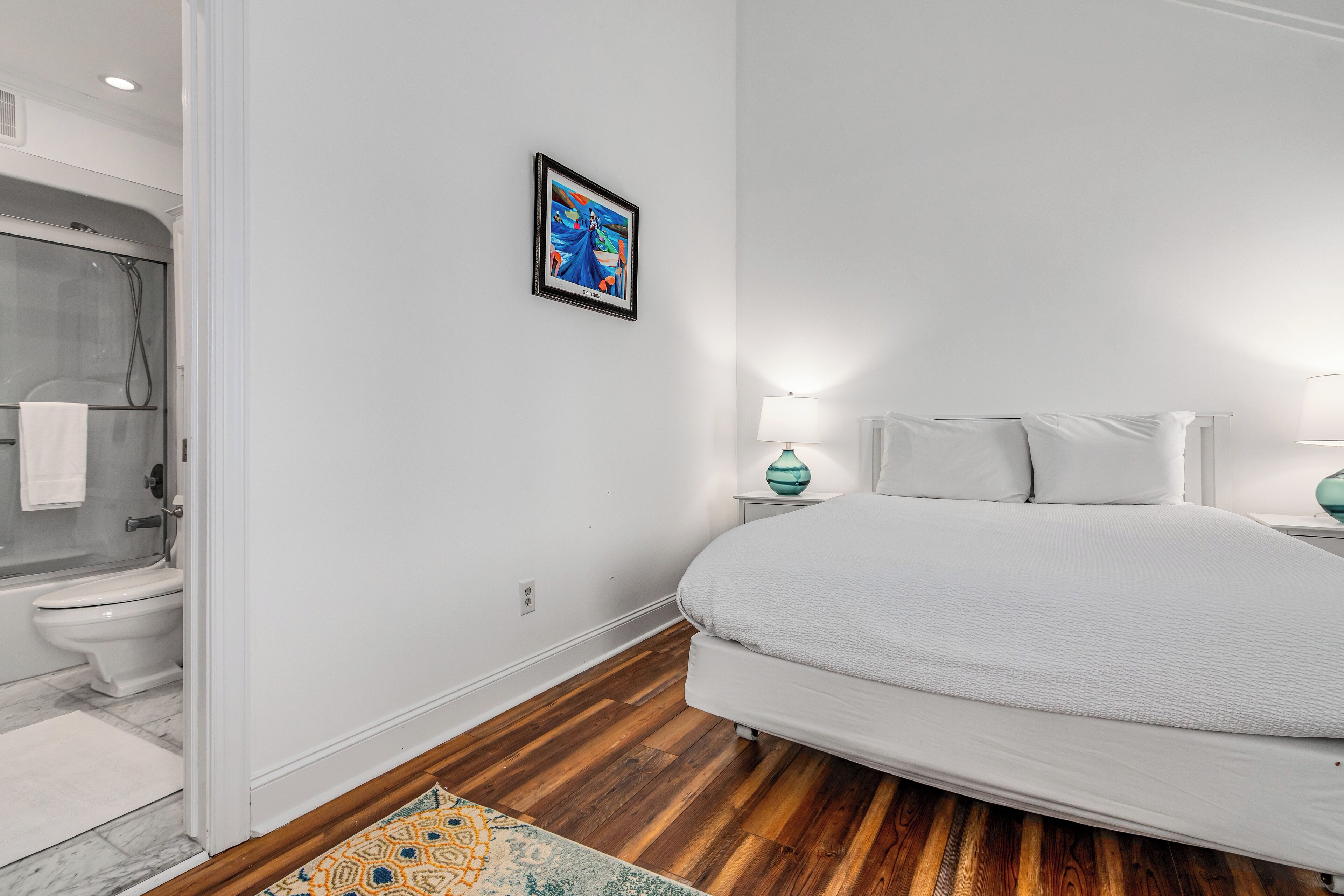 A bedroom with a large bed, a nightstand with a lamp, a framed picture on the wall, and a shower area.