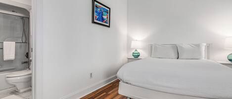 A bedroom with a large bed, a nightstand with a lamp, a framed picture on the wall, and a shower area.