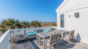 Una terraza de madera con mesa y sillas, baranda blanca y vista a la playa.