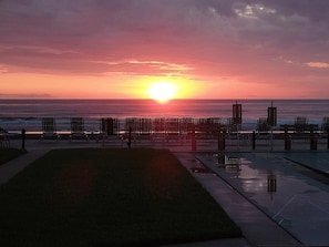 Vista del atardecer desde una zona de estar frente a la playa, con hileras de sillas y césped al frente.