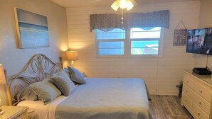 A bedroom with a bed, bedside tables, a dresser, a TV, a window with curtains, and a painting on the wall. - 202 - Adorable 1 bedroom unit - sleeps 6 (New Smyrna Beach)