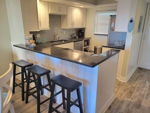 A kitchen with a central island, bar stools, and a window with blinds. - 202 - Adorable 1 bedroom unit - sleeps 6 (New Smyrna Beach)