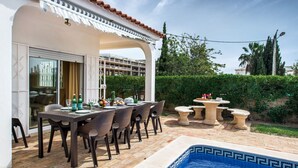 Un espace repas aménagé au bord de la piscine, avec une table dressée pour six personnes, entourée de chaises, et une petite table ronde sur laquelle sont posés quelques objets.