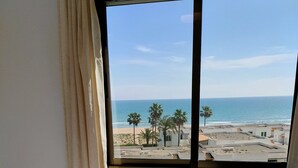 Een kamer met een bed en een raam met uitzicht op een strand met palmbomen en gebouwen.
