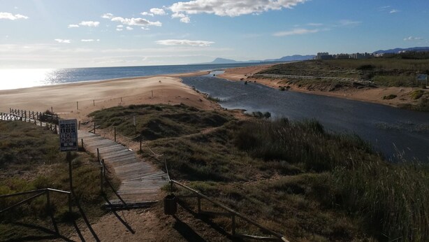 Un paysage côtier comprenant une plage, une rivière et une promenade en bois menant à un panneau indicateur.
