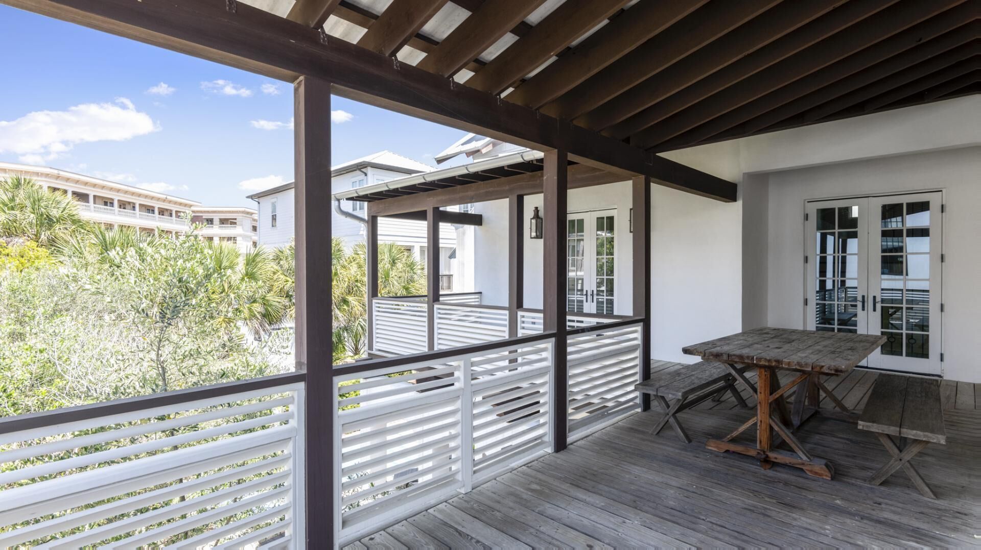 Une terrasse extérieure couverte, dotée d’une table et de bancs en bois, de rampes blanches et offrant une vue sur les bâtiments et la végétation environnants.