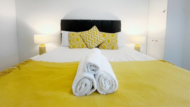 A hotel room with a bed, two bedside lamps, a headboard, and a rolled towel on the bed. - Anstey Heights - Apartment 4 - Hopewell (Easton)