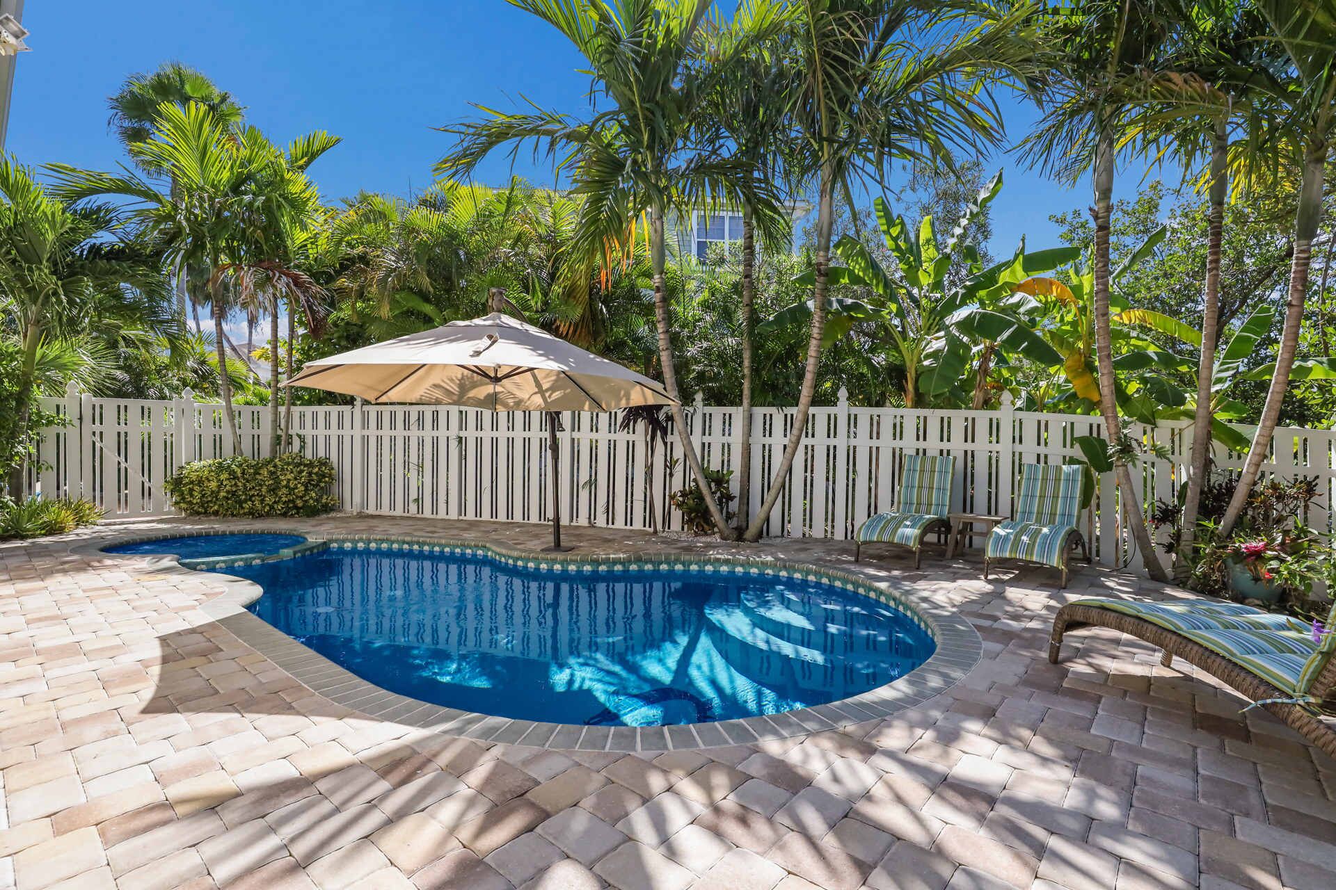 A swimming pool with a curved edge, surrounded by a white picket fence, palm trees, and a covered seating area with striped cushions.
