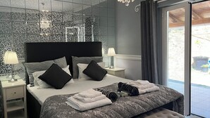 A modern bedroom with a large bed, a chandelier, and a view of the outdoors. - Villa Alexakis (corfu)
