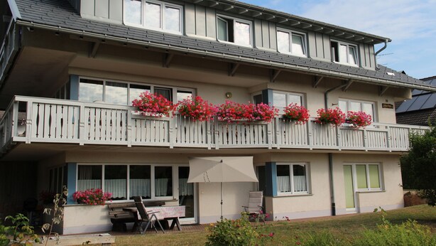 Ein zweistöckiges Gebäude mit einem an Blumen bepflanzten Balkon sowie einer überdachten Terrasse und einer sichtbaren Tür.